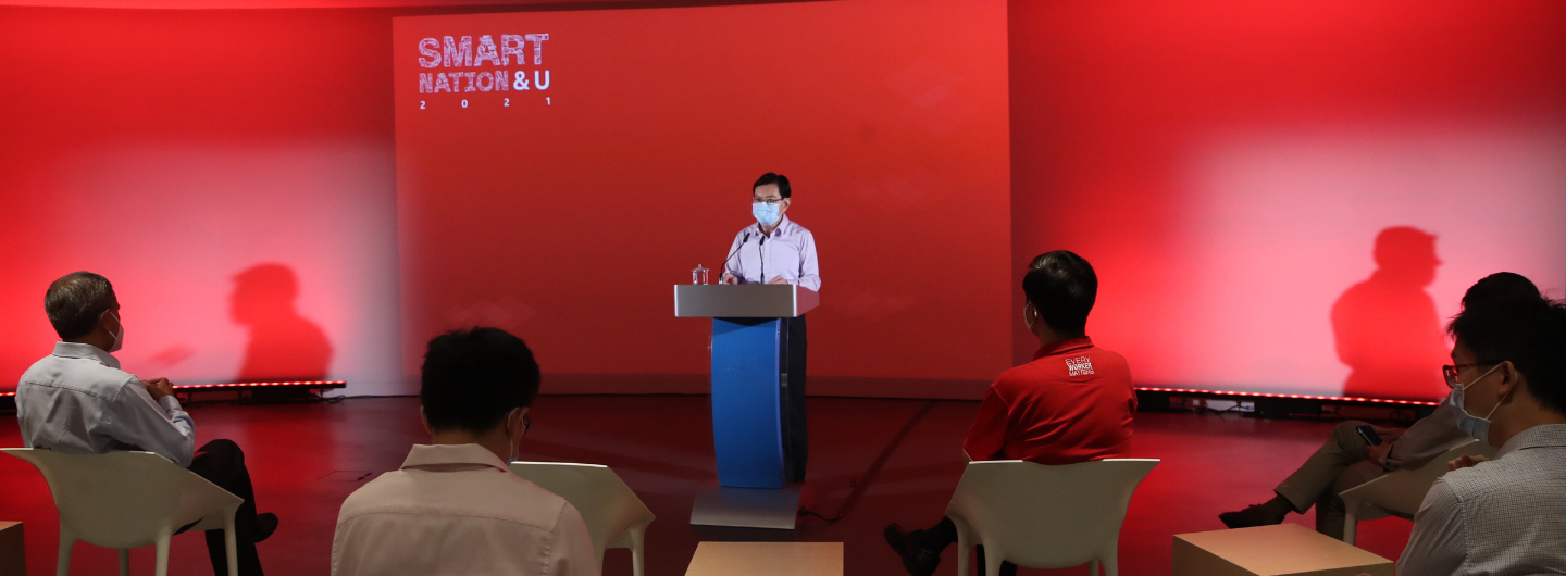 Speaker at podium, "SMART NATION & U" backdrop, audience seated in red-lit room.
